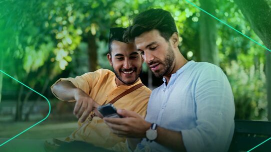 Dois homens sorrindo e usando smartphone em parque