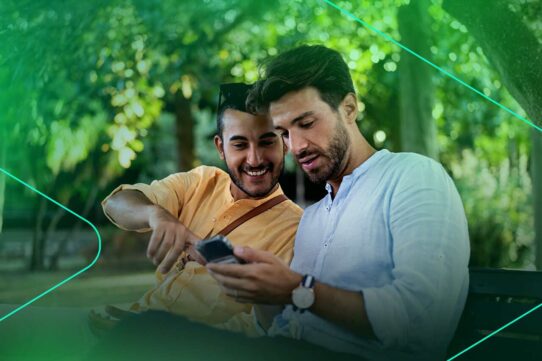 Dois homens sorrindo e usando smartphone em parque