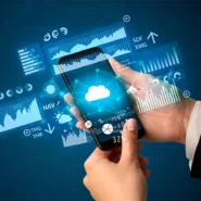 Mão segura smartphone exibindo ícone de nuvem cercado por gráficos digitais e análises financeiras azuis.