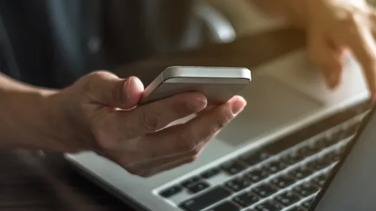 Pessoa interagindo em um notebook com o celular na mão