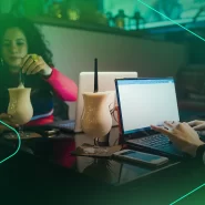 Duas pessoas trabalhando em laptops com bebidas ao fundo.