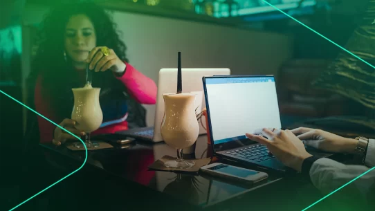 Duas pessoas trabalhando em laptops com bebidas ao fundo.