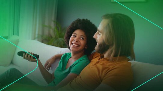 Casal sorrindo enquanto assiste TV no sofá.