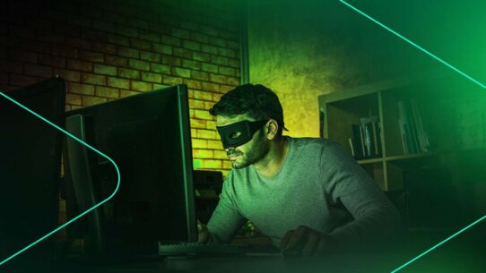 Homem com máscara usando computador em ambiente escuro.