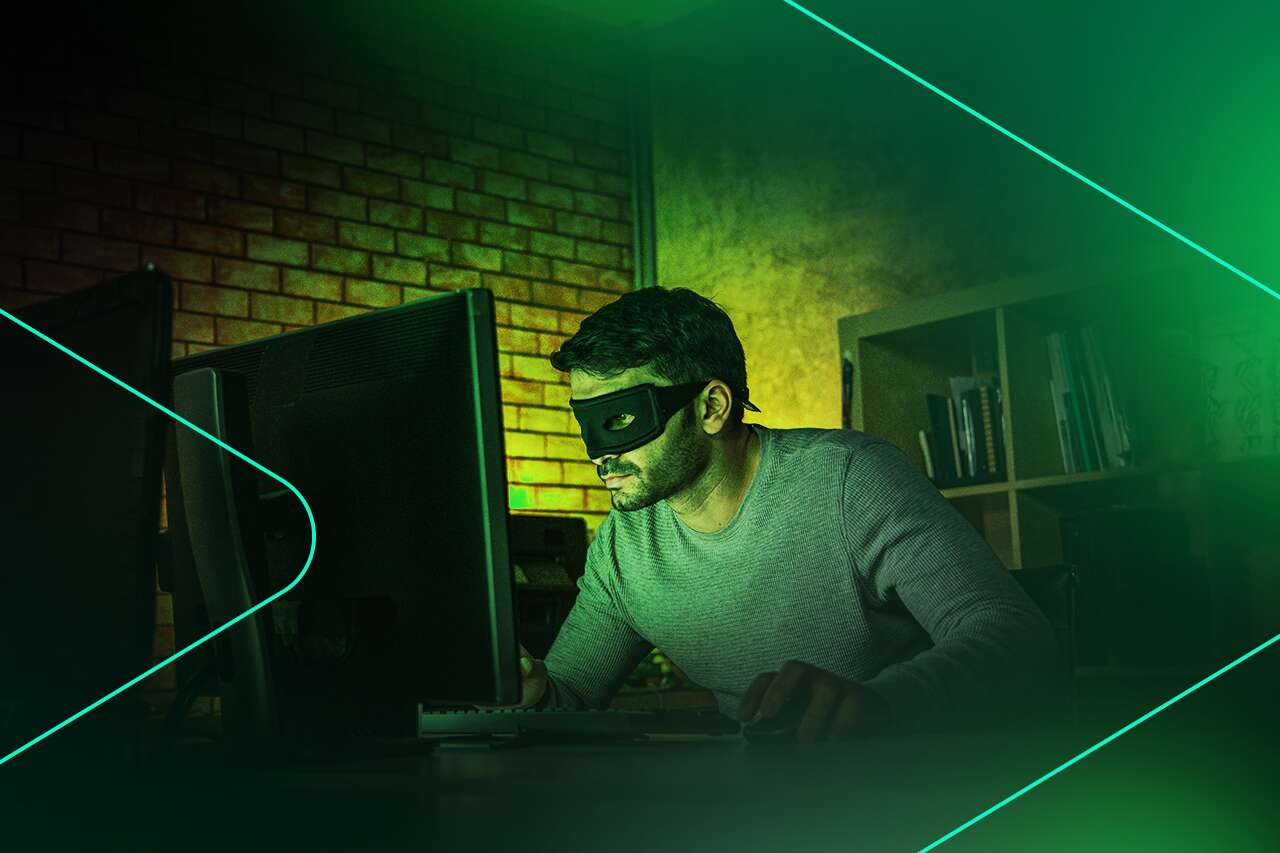 Homem com máscara usando computador em ambiente escuro.