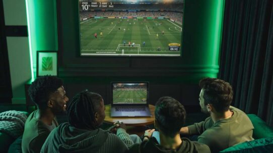 Grupo assistindo futebol em casa com laptop e TV.