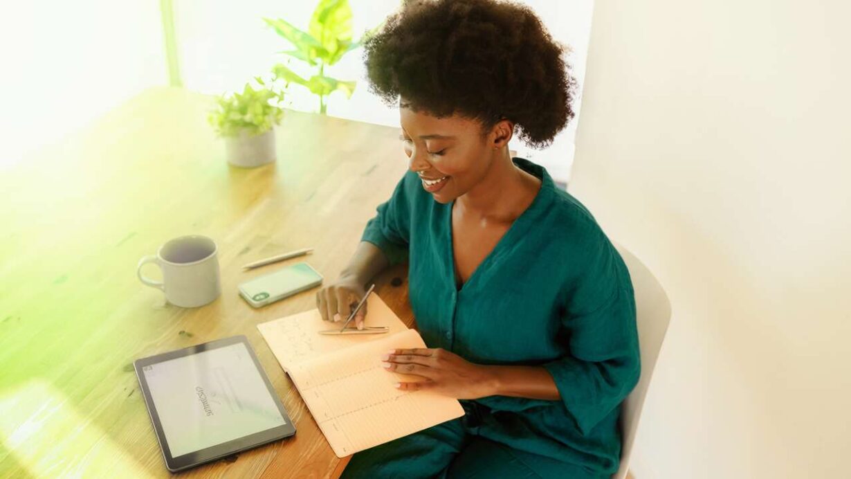 Mulher sorridente escrevendo em caderno na mesa.