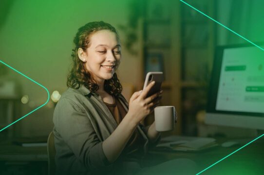 Jovem sorrindo enquanto usa o celular e segura uma xícara.