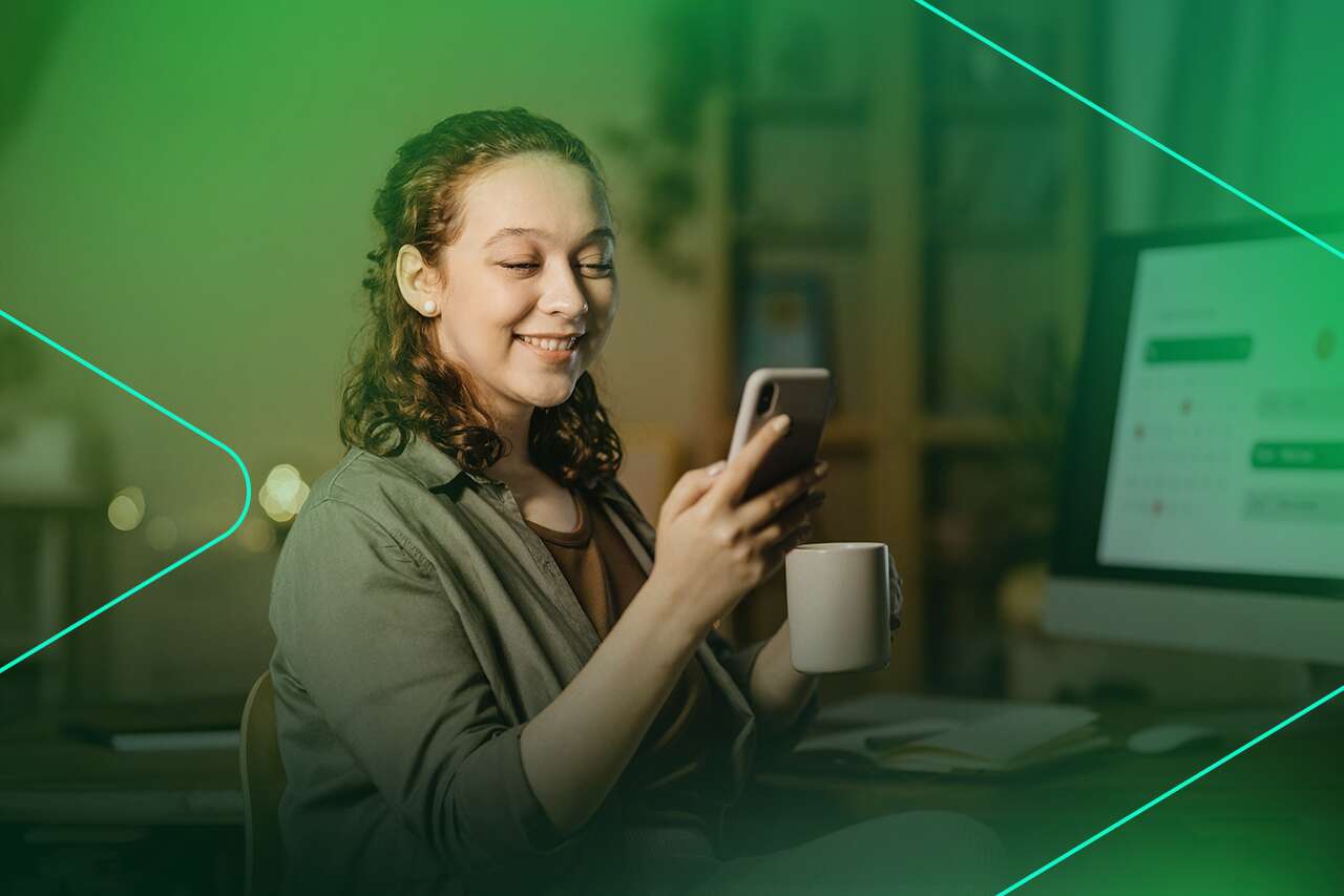 Jovem sorrindo enquanto usa o celular e segura uma xícara.