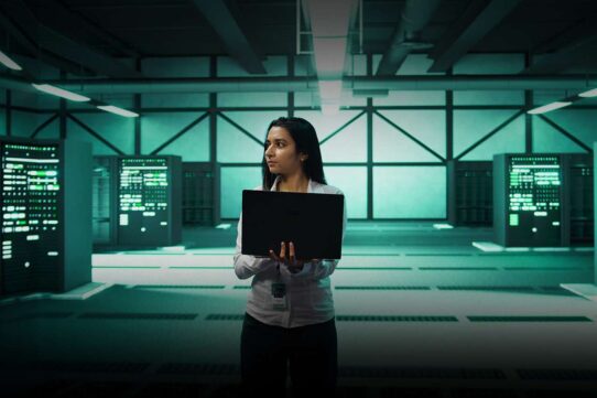 Mulher segura laptop em sala de servidores modernos.