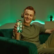 Homem sorridente com fones, usando celular no sofá.