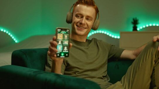 Homem sorridente com fones, usando celular no sofá.
