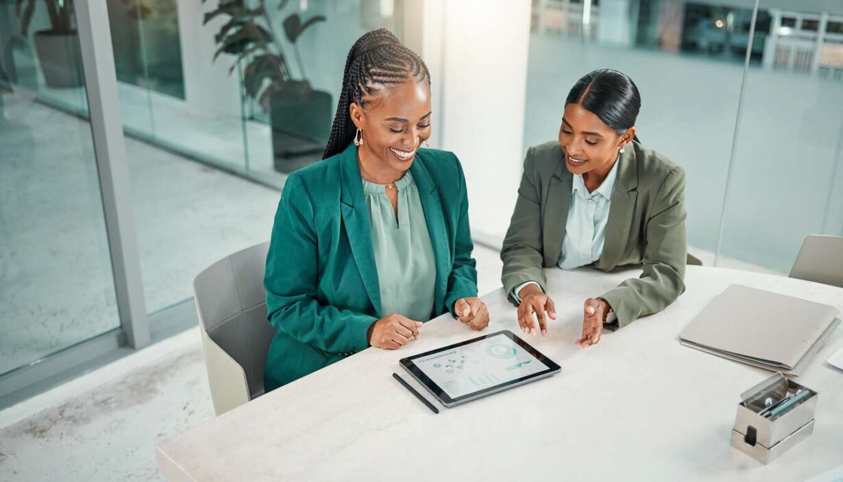 Duas mulheres analisando dados em um tablet em escritório moderno.
