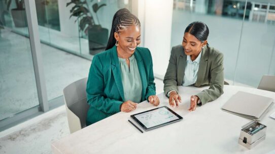 Duas mulheres analisando dados em um tablet em escritório moderno.